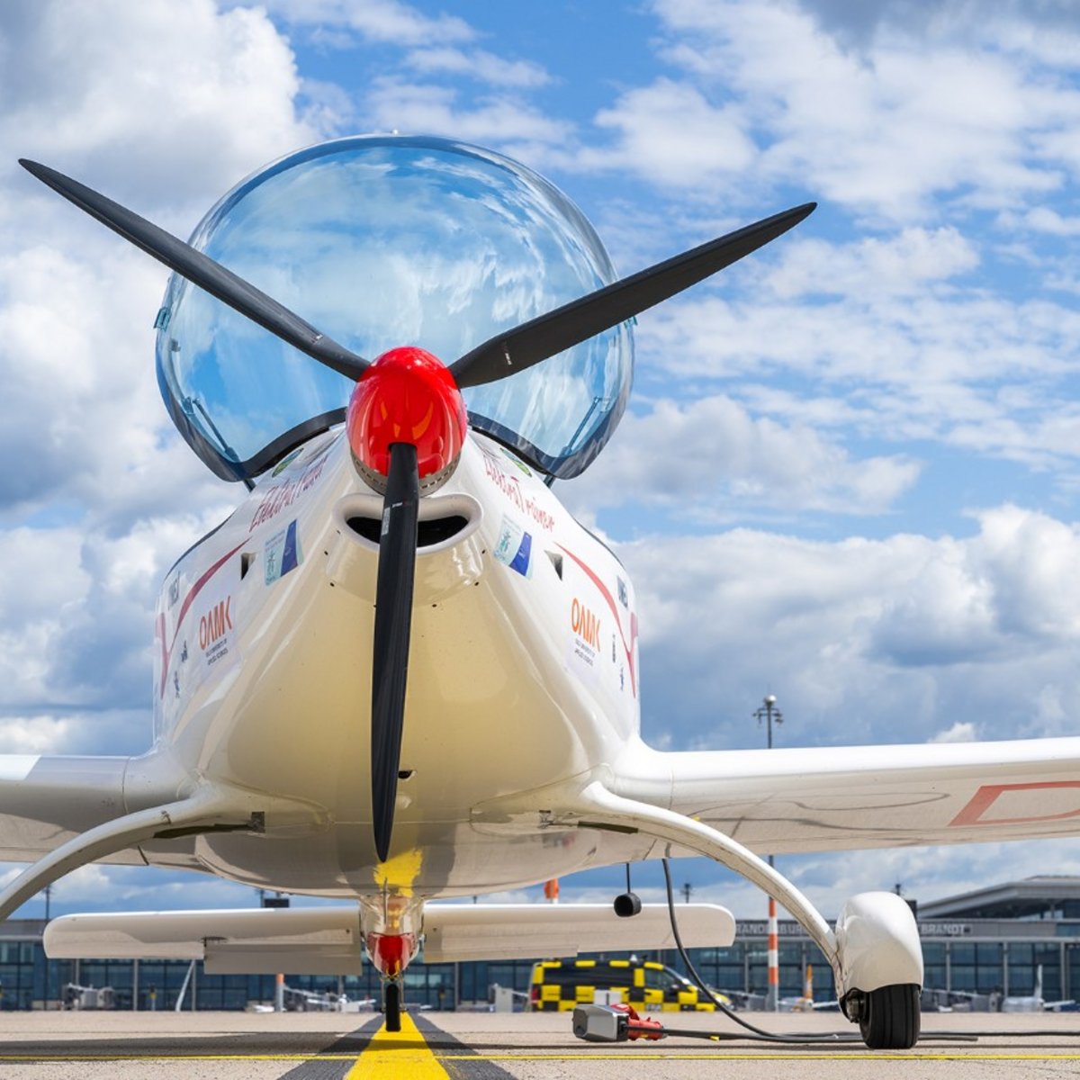 Blick von unten auf den vollelektrischen „Elektra Trainer“: Der Propeller und die Frontpartie des Flugzeugs sind frontal zu sehen, darüber zeichnen sich Rumpf und Tragflächen gegen den Himmel ab.