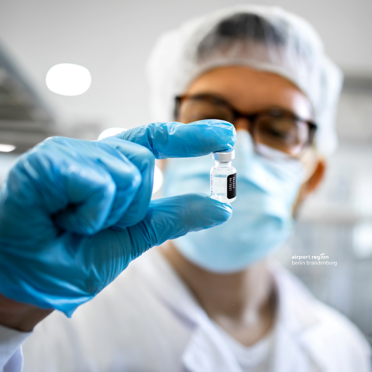 Bild zeigt einen Forscher mit seiner Forschungsuniform: einemweißem Kittel, Mundschutz und Haube. Im Vordergrund sieht man seine rechte Hand, die einen blauen Handschuh trägt. Zwischen Dauemn und Zeigefinger zeigt der Mann ein kleines Fläschchen mit einer klaren Flüssigkeit - in diesem Fall ein Impfstoff.