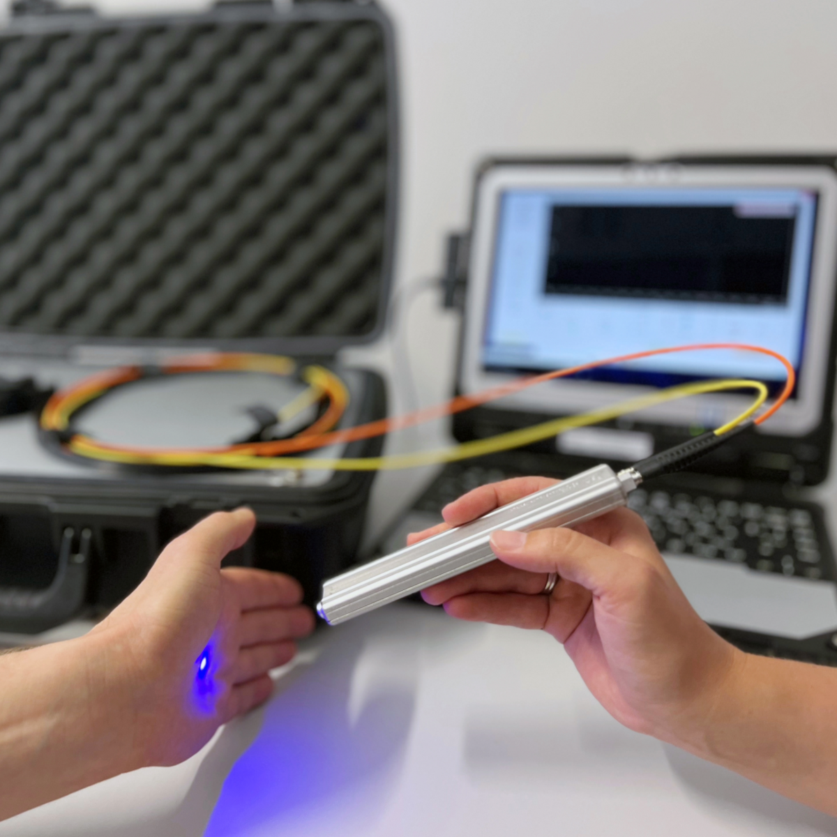 Bild zeigt zwei Hände, die ein optisches Gerät vor einem Messcomputer halten. Das Gerät erzeugt auf der Haut einen blauen Lichtpunkt und misst damit bestimmte Werte in der Haut.