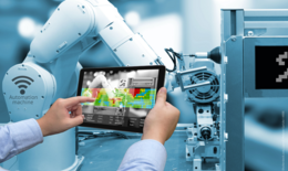 Bild zeigt einen Roboterarm und zwei menschliche Hände, die ein Bedienpanel halten und den Roboterarm steuern. Das ganze befindet sich in einer fabrikähnlichen Umgebung. Es greift die Robotik als Thema auf, welche im Artikeltext eine Rolle spielt.