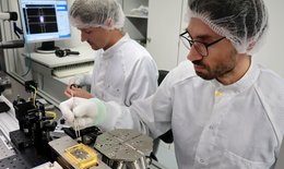 Zwei Forscher mit Haarnetz, Handschuhen und weißem Kittel bearbeiten im Labor Technikelemente.