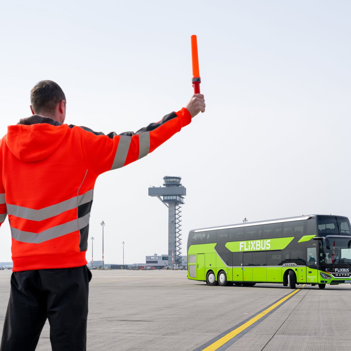 Symbolisch hält ein Fluglotse die Richtungsstäbe in Richtung Flixbus vor dem Tower am Berliner Flughafen