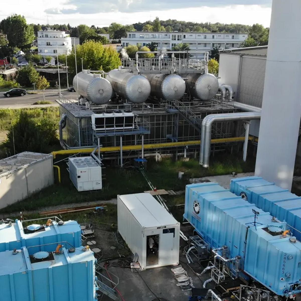 Übersicht über die Bohrstelle in Berlin Adlershof, die sich auf dem Gelände der BTB GmbH befindet. Das aus dem Hettang-Sandsteinreservoir geförderte Formationswasser wird während der Pull-Phasen in den blauen Flüssigkeitstanks gespeichert. Helle, silberne, isolierte Rohre leiten die heiße Flüssigkeit vom Wärmetauscher (der sich zwischen dem weißen Laborcontainer und dem Flüssigkeitstank befindet) über die Flüssigkeitsüberwachungs- und -messeinheit zum Forschungsbohrloch.