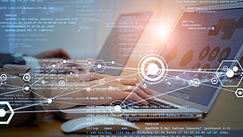 Bild zeigt zwei Laptops im Hintergrund, im Vordergrund ein schematische Darstellung eines Netzwerks, Programmiercodeelemente sind sichtbar. Das Bild ist ein Synonym für Digitalisierung.