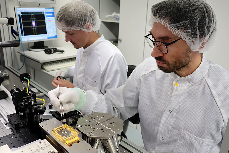 Zwei Forscher mit Haarnetz, Handschuhen und weißem Kittel bearbeiten im Labor Technikelemente.